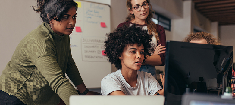 How to Attract Women in Technology: a diverse group of women gathered around a monitor focused on their work.